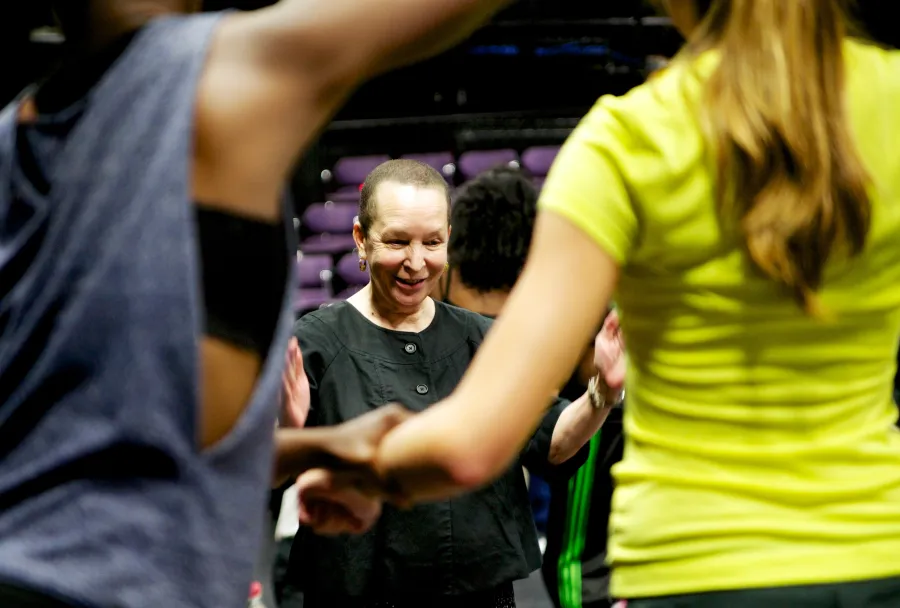 A woman in black leads a group activity, smiling, with participants in bright workout clothes visible in the foreground.