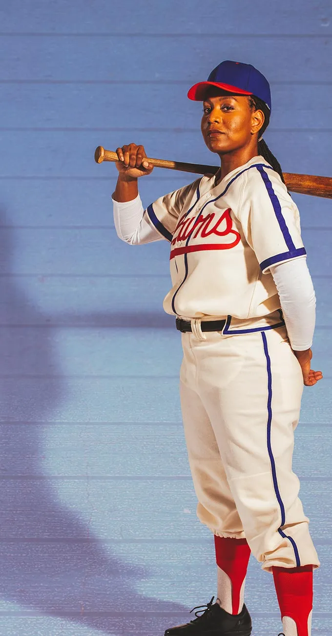 A woman in a vintage baseball uniform holds a bat over her shoulder, standing on a blue-tinted surface and looking confidently at the camera.