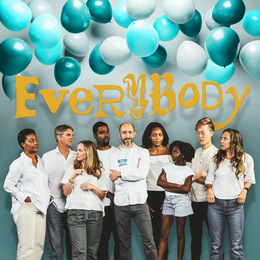 A diverse group of people in white clothing stands under blue and white balloons with the word "EVERYBODY" in large yellow letters on the wall behind them.