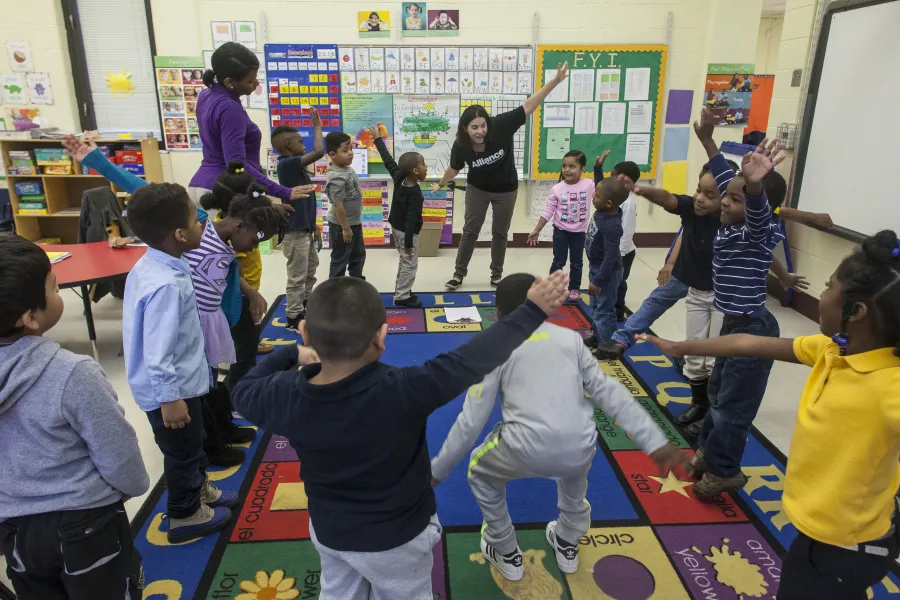 A group of young children and two adults stand in a classroom, participating in an activity together while forming a circle on an alphabet rug.