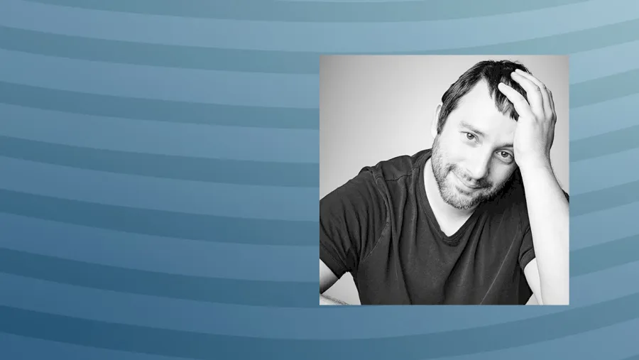 A man with a beard and short hair, wearing a dark t-shirt, sits with one hand on his head against a striped blue background.