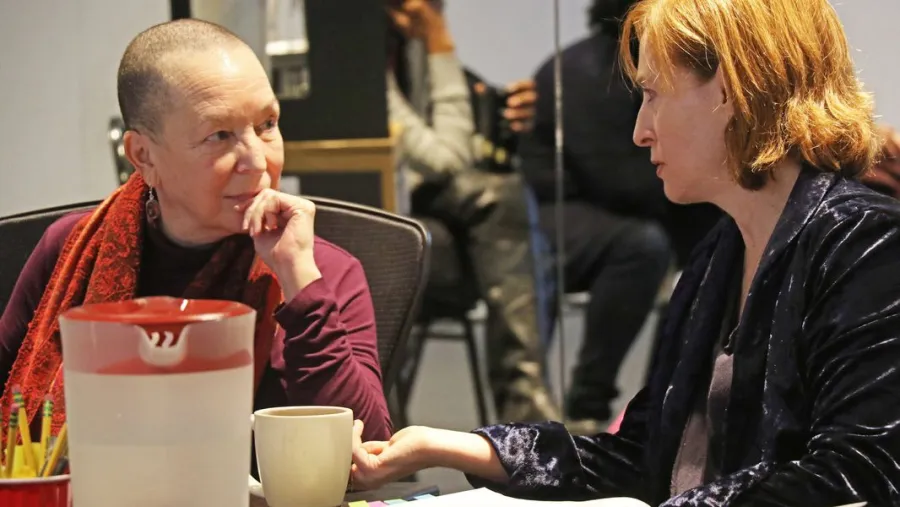 Two women sit at a table engaged in conversation. One has a shaved head and wears a red scarf, the other has short blonde hair and wears a dark jacket. A pitcher and mug are on the table.