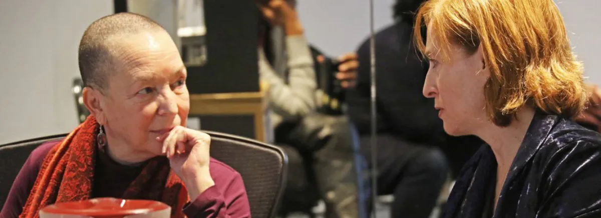 Two women sit facing each other and engage in a thoughtful conversation indoors; one has short hair, the other has light brown hair.