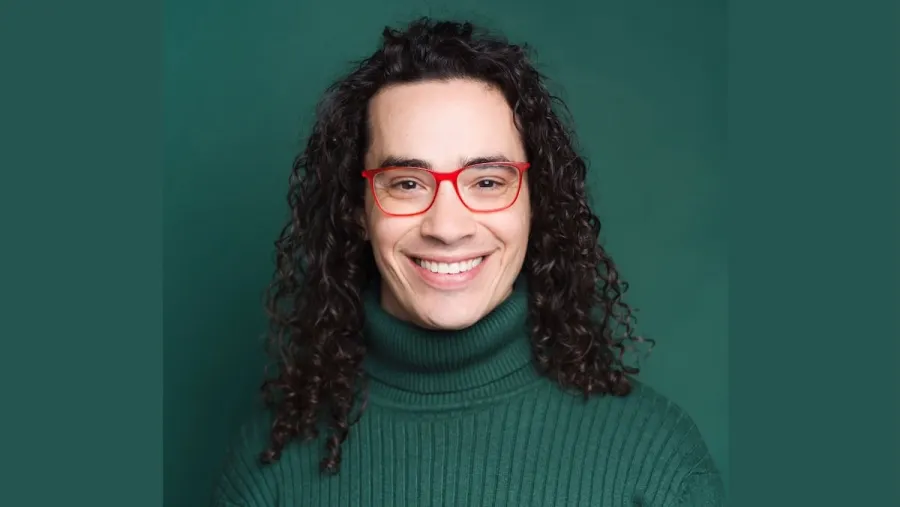 Person with long curly hair wearing red glasses and a green turtleneck sweater, smiling in front of a green background.
