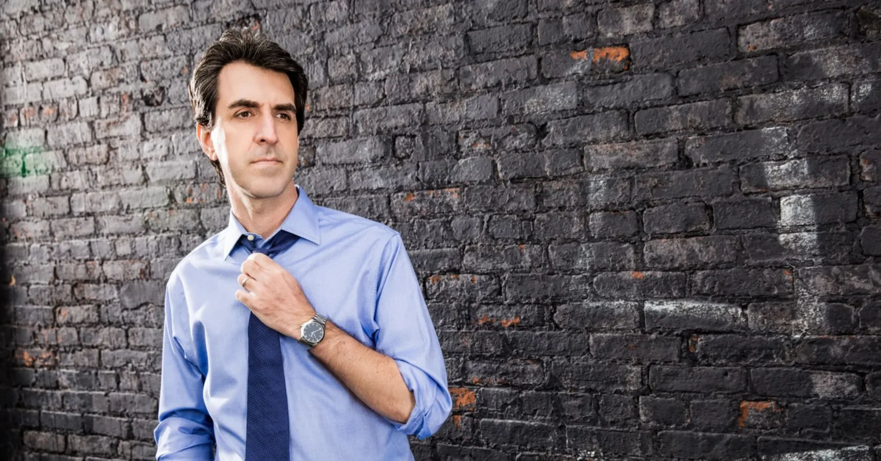 A man in a blue dress shirt adjusts his tie while standing in front of a weathered brick wall.