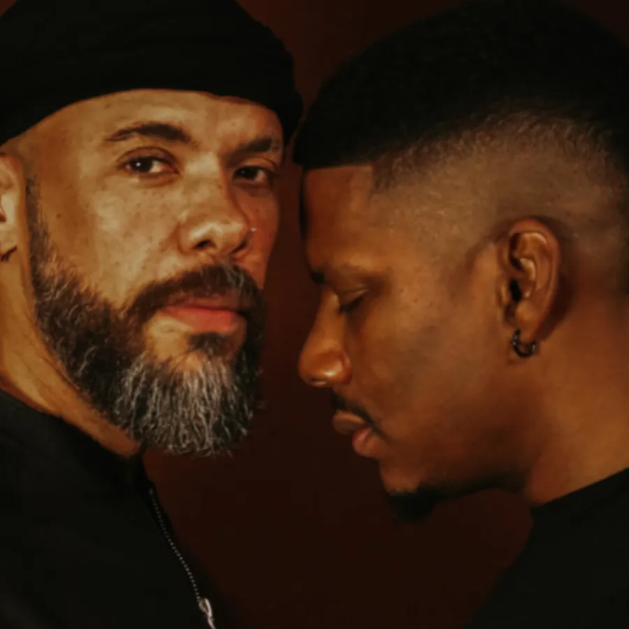 Two men stand close together against a dark background; one faces the camera with a neutral expression, while the other faces sideways with eyes closed.