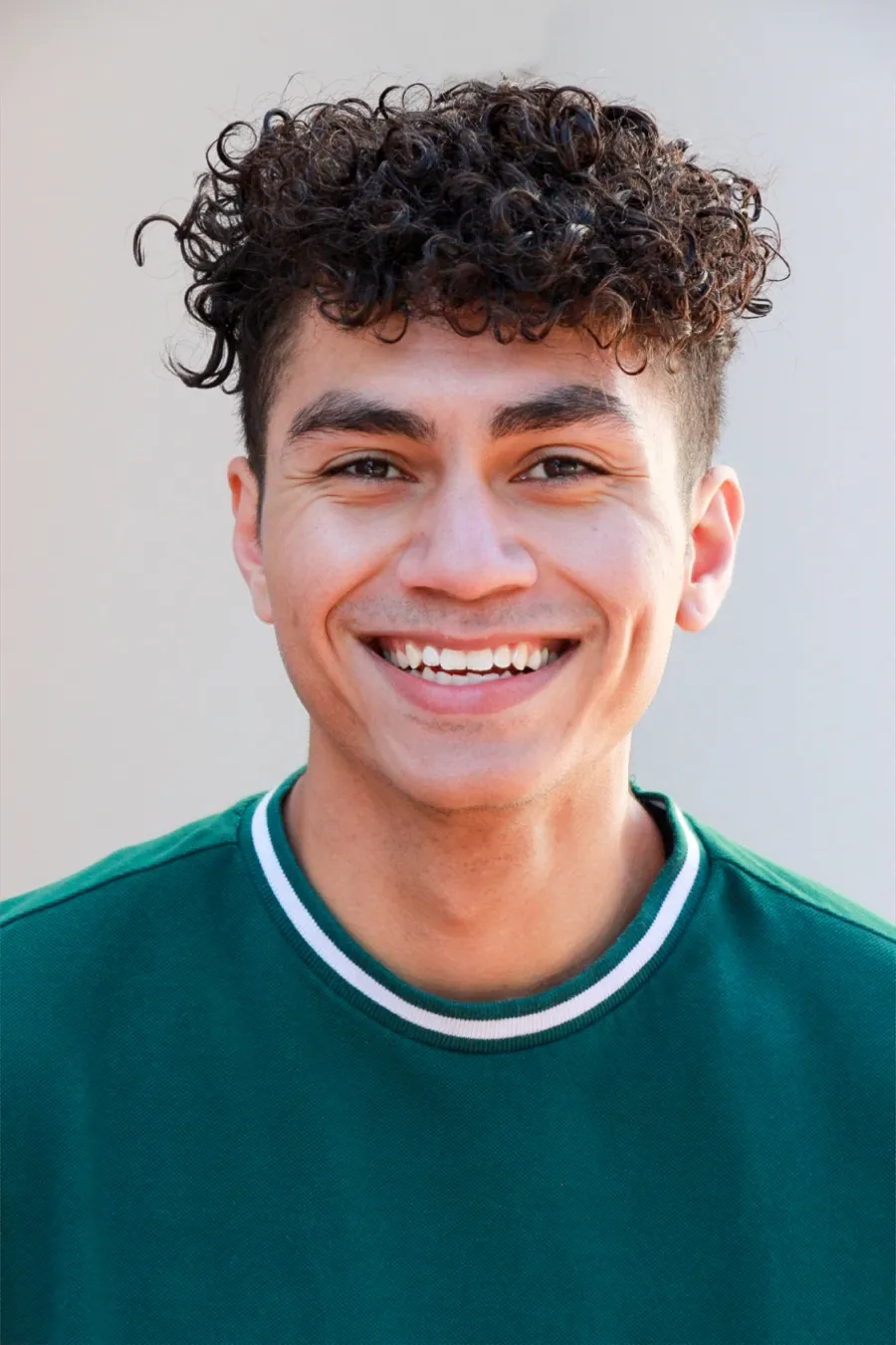 Young man with curly hair wearing a green sweatshirt smiles at the camera against a plain light background.