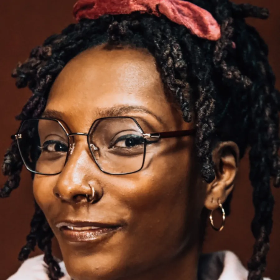 A person with glasses, a nose ring, hoop earring, and dreadlocks tied with a red scrunchie, smiles slightly while looking at the camera.