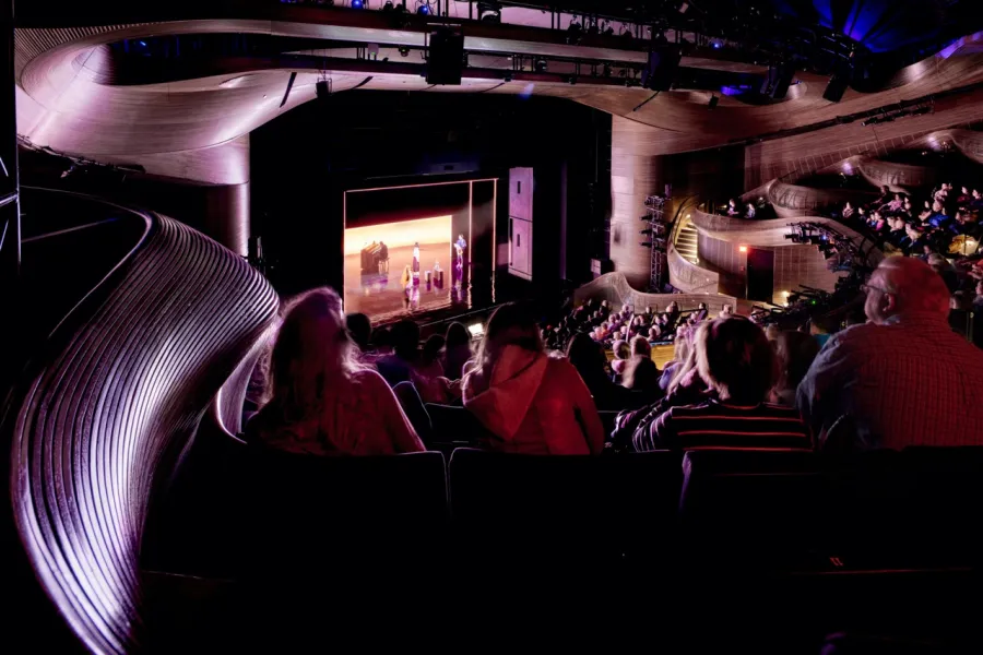 Audience seated in a modern, dimly lit theater watching a live stage performance with illuminated actors on stage.