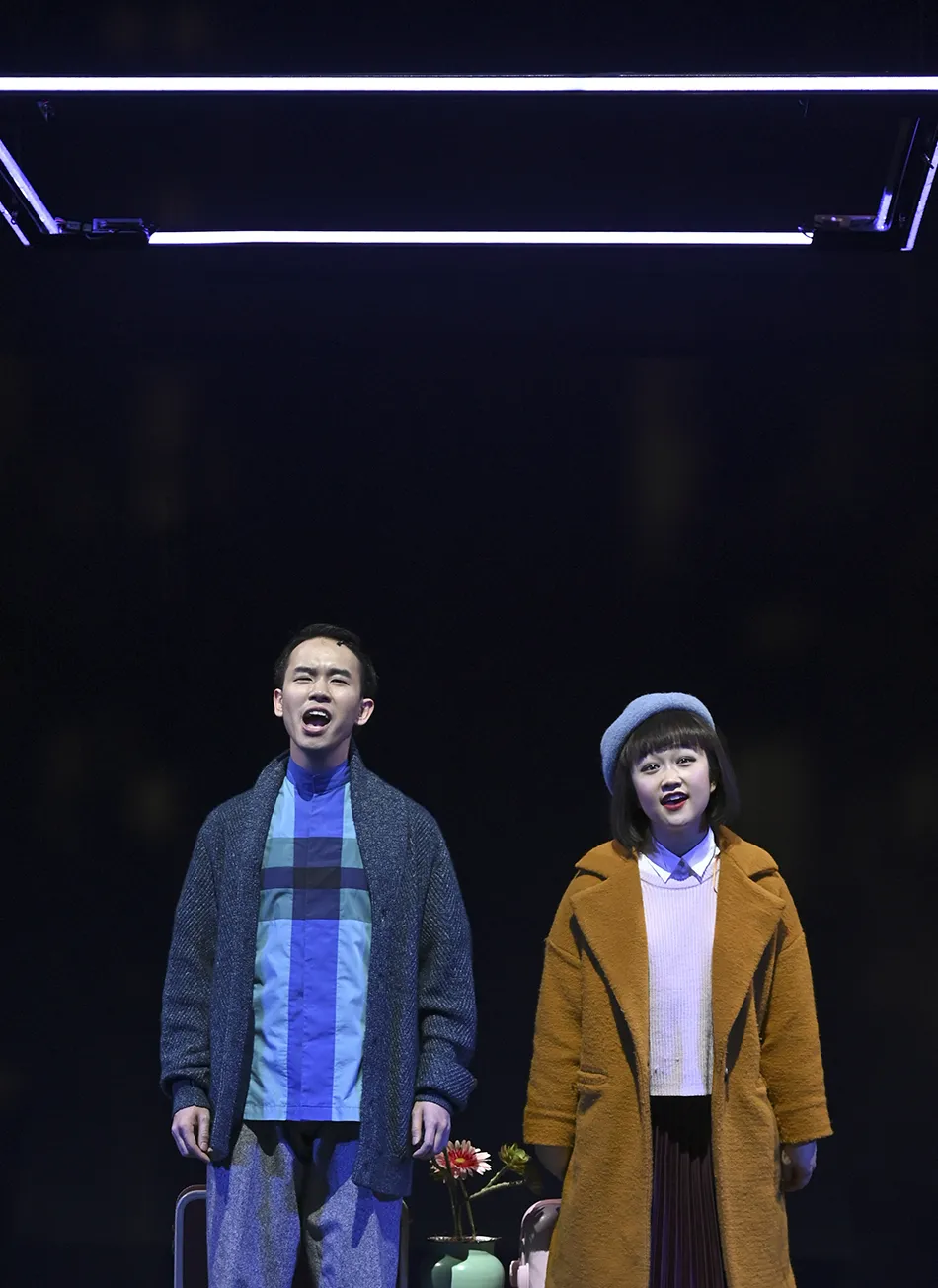 Two people stand under a square frame of lights on a dark stage, wearing winter coats; a small table with flowers and vases is between them.