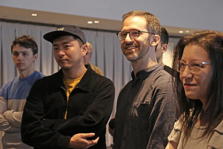 A group of four people stand indoors, facing forward. Two are smiling, while two have neutral expressions. A curtain is visible in the background.