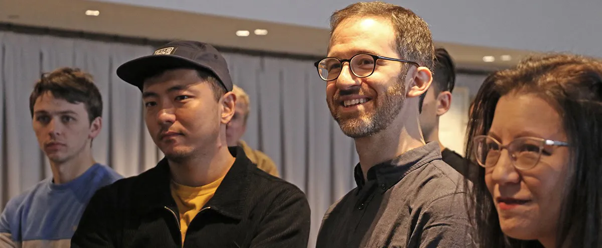 Four adults stand indoors, facing forward. Three men and one woman are visible, all wearing casual clothing and glasses. The background features curtains and soft overhead lighting.