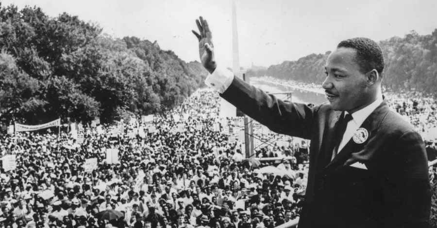 A man in a suit waves to a large crowd gathered outdoors on a tree-lined street, with the Washington Monument visible in the distance.