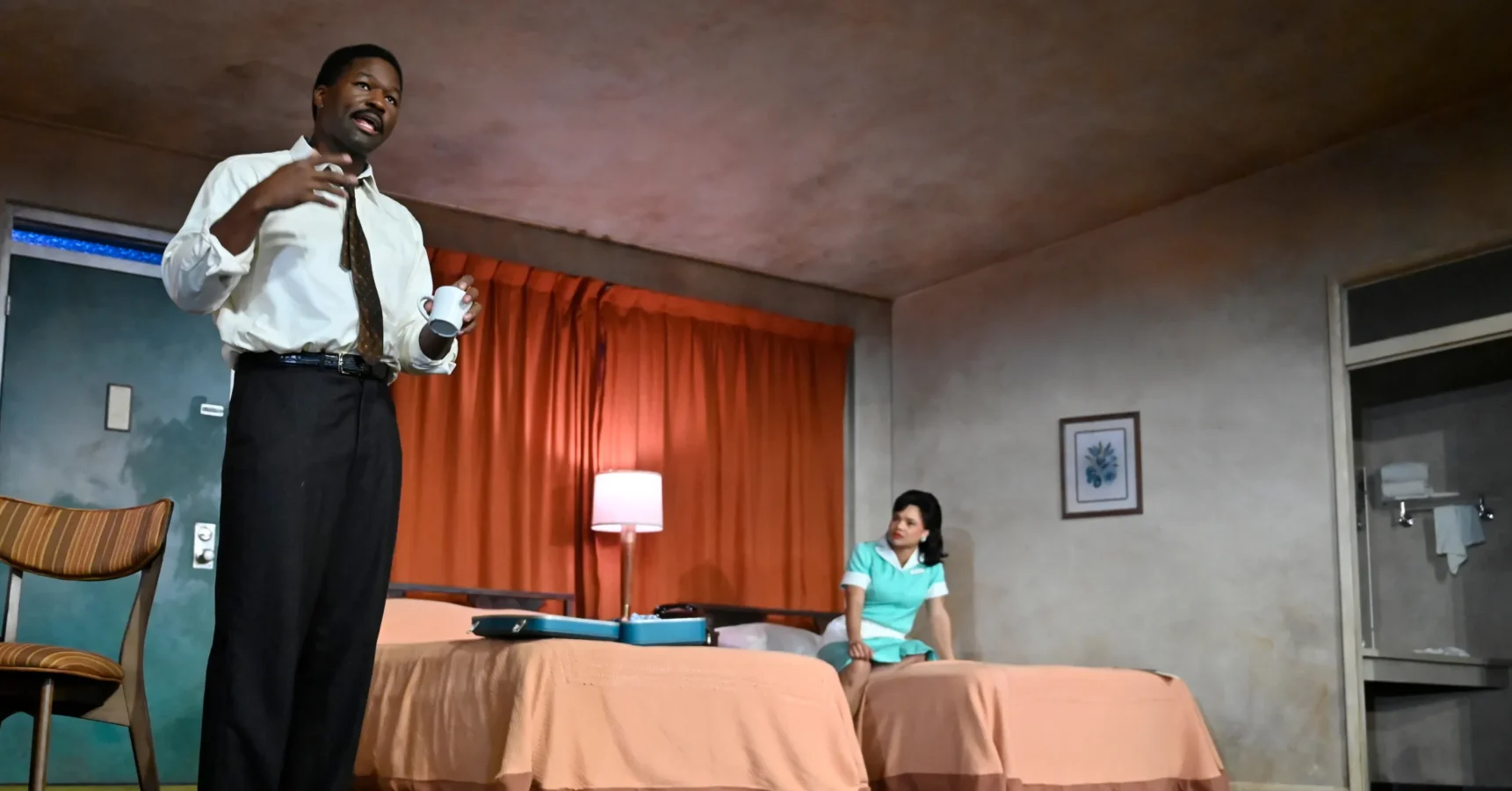 A man in a dress shirt and tie holds a mug while speaking; a woman in a green dress sits on a bed in a sparsely furnished room with orange curtains and peach bedspreads.