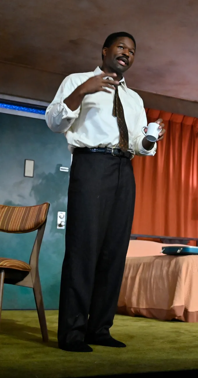 A man in a white shirt, dark pants, and tie stands near a chair, holding a cup and gesturing with his other hand in a room with orange curtains.