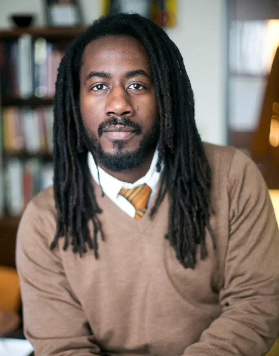 A man with long dreadlocks, a brown sweater, and a striped tie sits indoors in front of a blurred bookshelf.