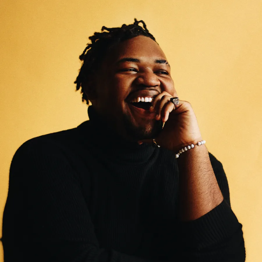 A person wearing a black turtleneck and beaded bracelet smiles and laughs with their hand touching their chin against a plain yellow background.