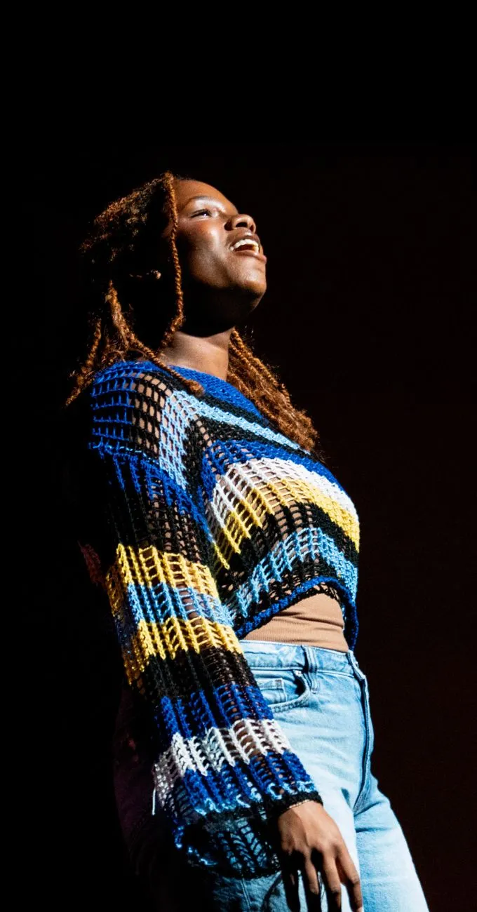A person wearing a colorful striped crochet sweater and jeans stands in a spotlight against a dark background, looking slightly upward with a neutral expression.