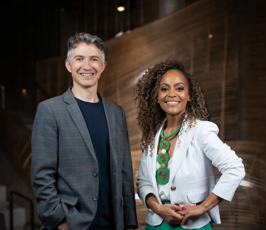 A man in a blazer and a woman in a white jacket stand side by side indoors, smiling at the camera against a modern wooden backdrop.