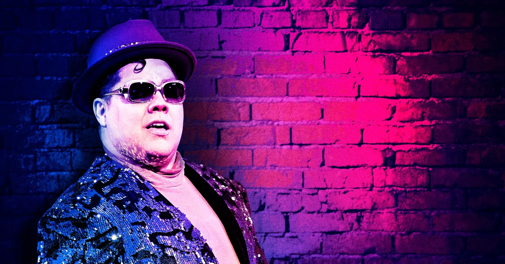 A person wearing sunglasses, a purple hat, and a sequined jacket stands in front of a brick wall lit with pink and blue lighting.
