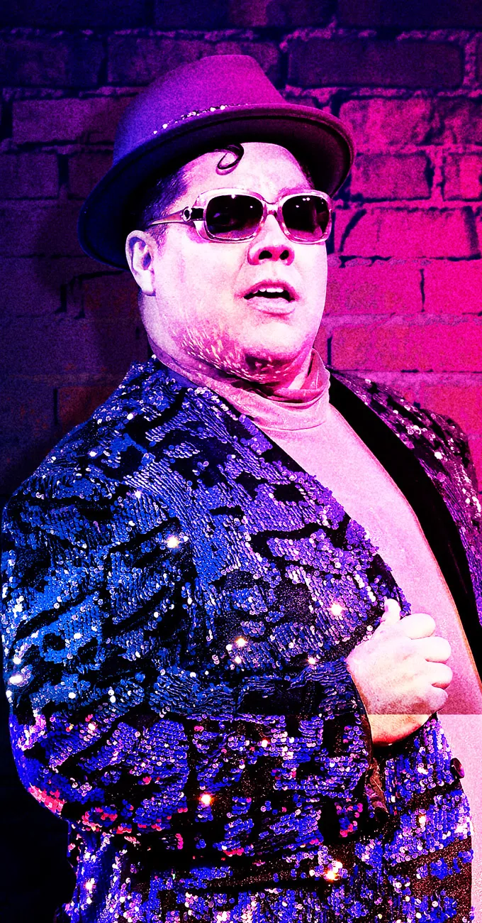 Person wearing a sequin jacket, sunglasses, and a hat stands in front of a brick wall under colorful pink and purple lighting.
