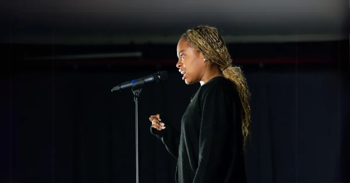 A person with long braided hair speaks into a microphone on a dimly lit stage, wearing a black sweater, against a dark background.