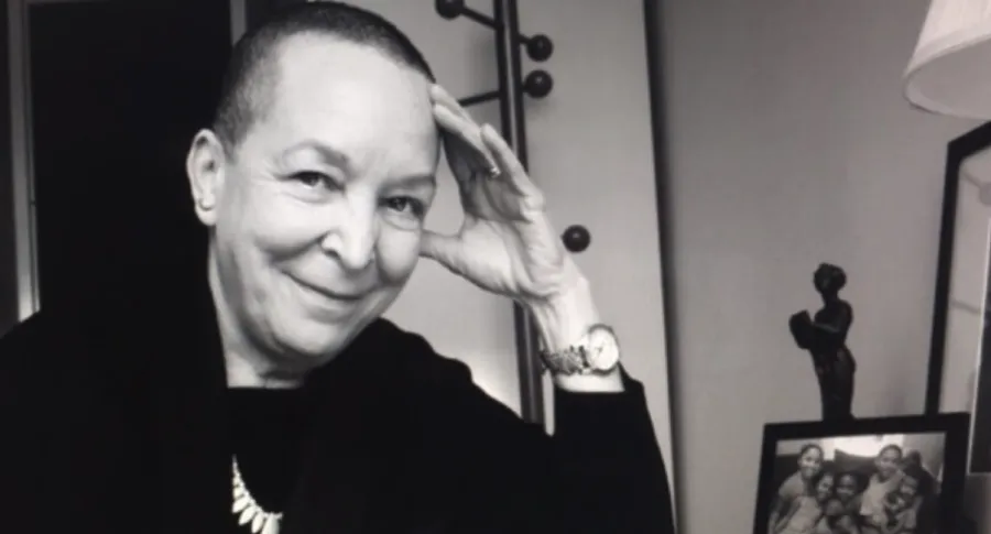 A person with short hair smiles at the camera, resting their head on their hand. In the background, a framed photo and a small sculpture are visible.