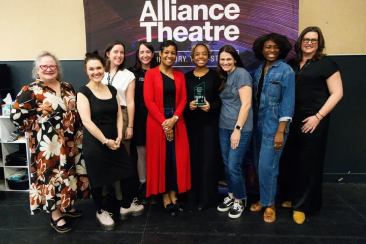 Nine women pose together indoors in front of an "Alliance Theatre" sign; one woman in the center holds a glass award.