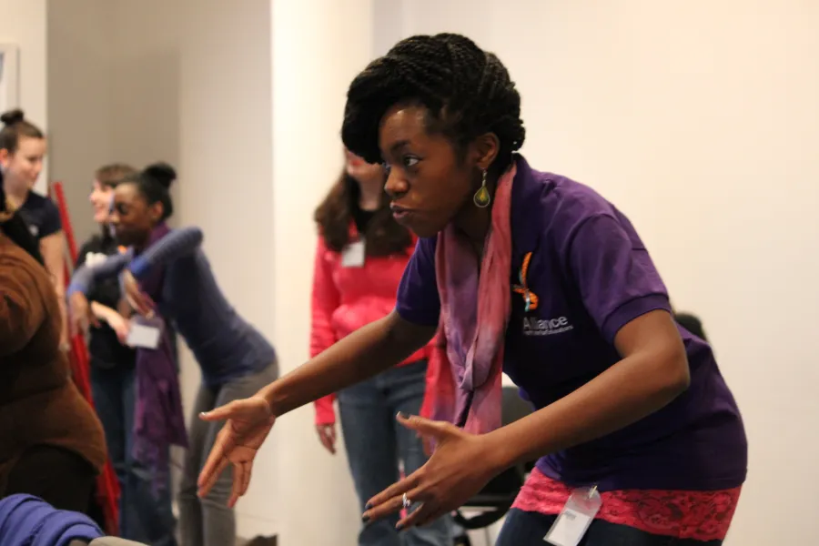 A woman in a purple shirt gestures with her hands while speaking to a group of people in a room; others are standing and interacting in the background.