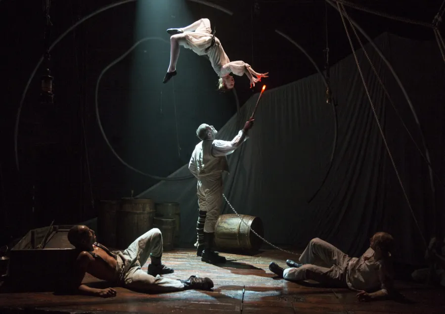 A person in white clothing is suspended in the air above a stage, while another person holds a lit torch beneath them. Two people are sitting on the floor, looking on.