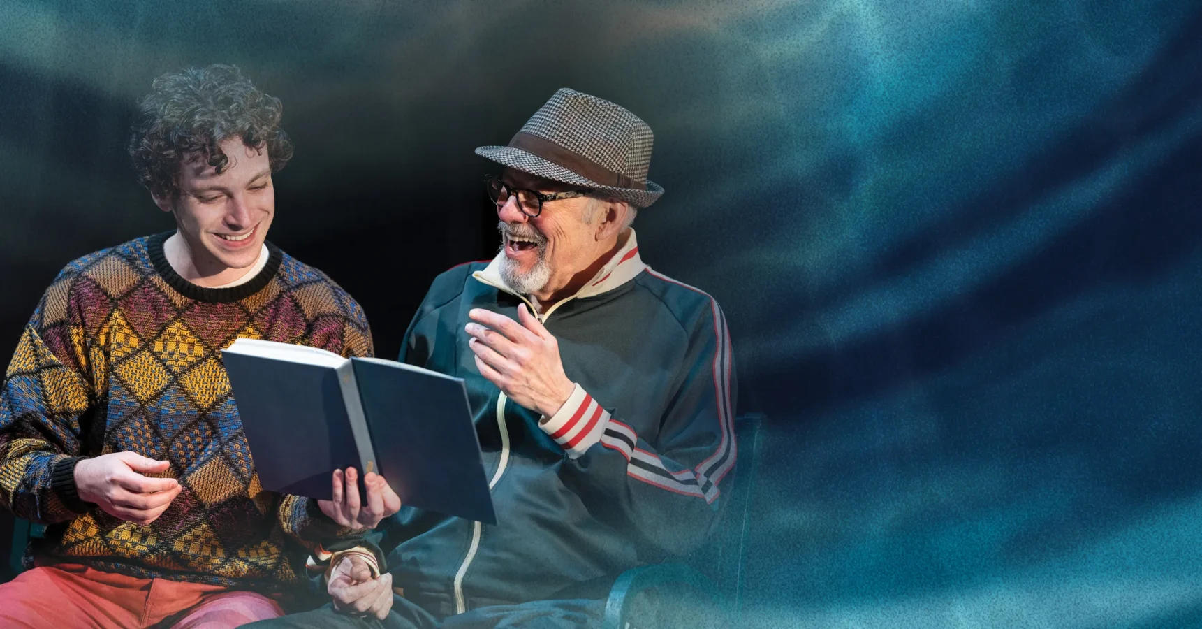 Two men sit together smiling while looking at an open book; one wears a colorful sweater, the other a hat and glasses, against a blue, softly lit background.
