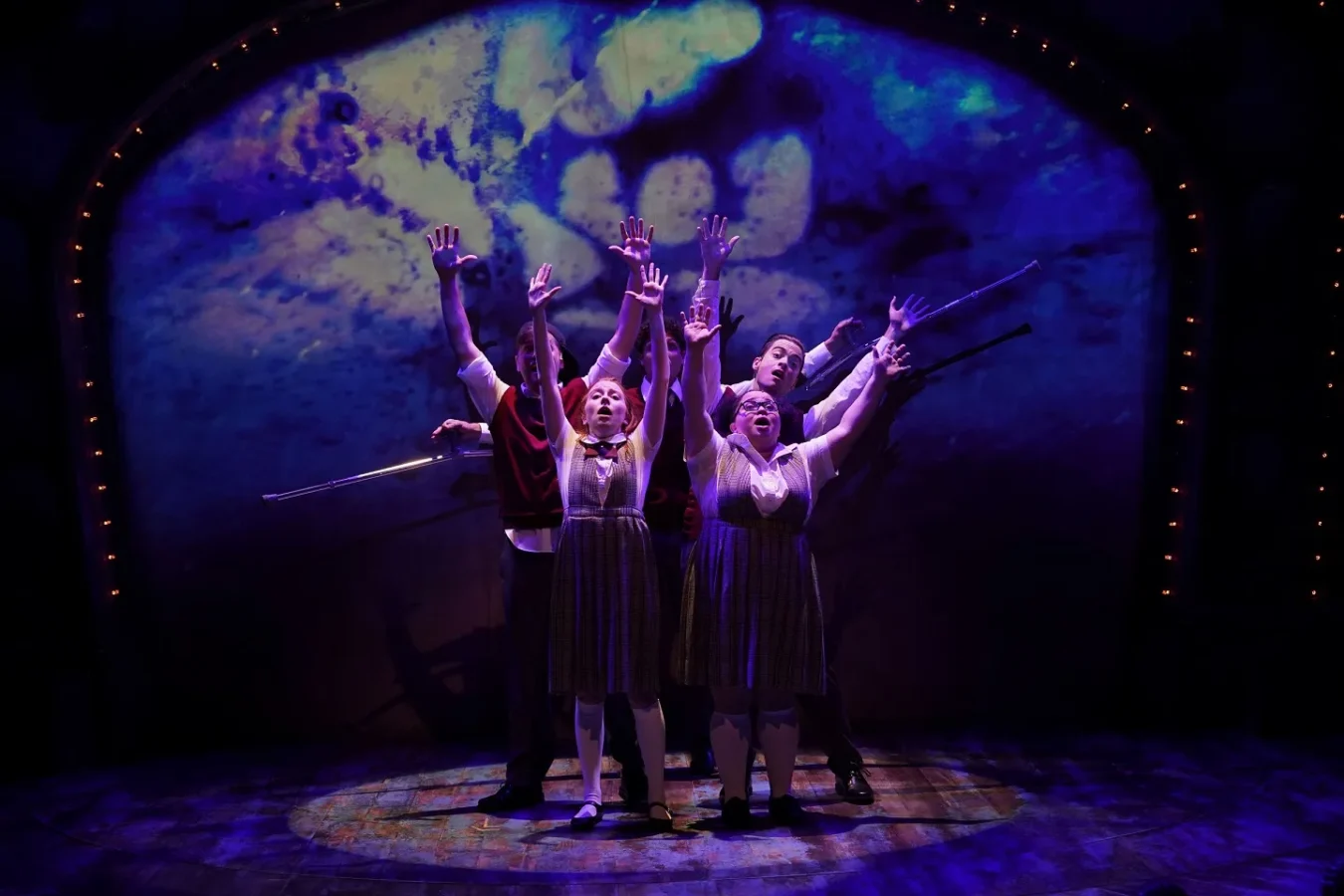 Five actors on stage, dressed in school uniforms, stand with arms raised dramatically under blue and purple stage lighting, with a swirling abstract backdrop behind them.