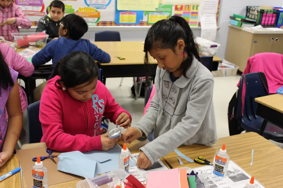 Two children work together on a craft project at a classroom table, using construction paper, glue, and other art supplies. Other students are visible in the background.