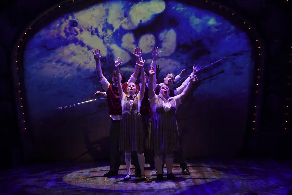 Five actors on stage reach upward with dramatic expressions, dressed in school uniforms, against a blue and white illuminated backdrop shaped like a cloud or a face.