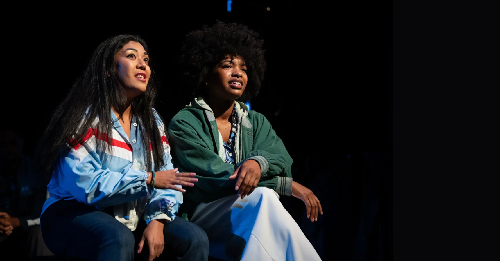 Two women sit side by side, attentively looking ahead; one wears a light blue jacket, the other a green jacket, both illuminated against a dark background.