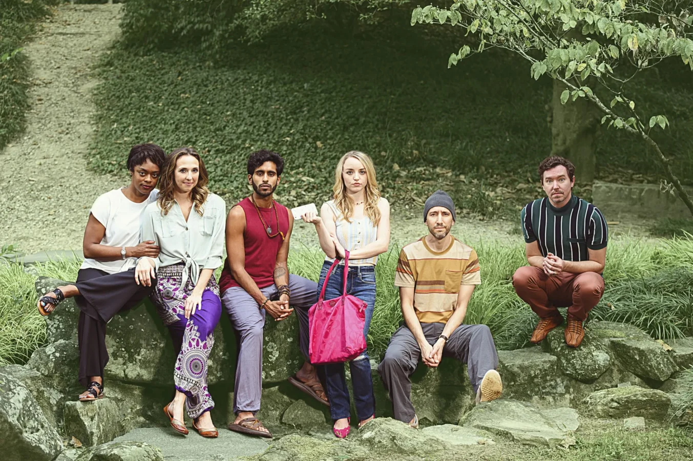 Six people sit and stand on rocks outdoors in a natural, green setting, posing for the camera with relaxed and casual expressions.