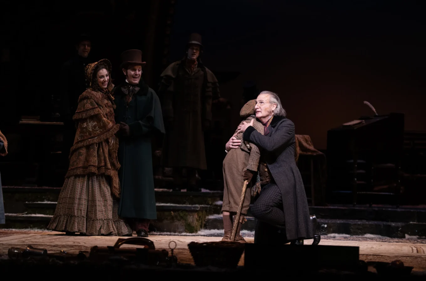 Four actors in period costumes perform on stage; one man kneels in the foreground, grasping another's leg, while two others stand together in the background.