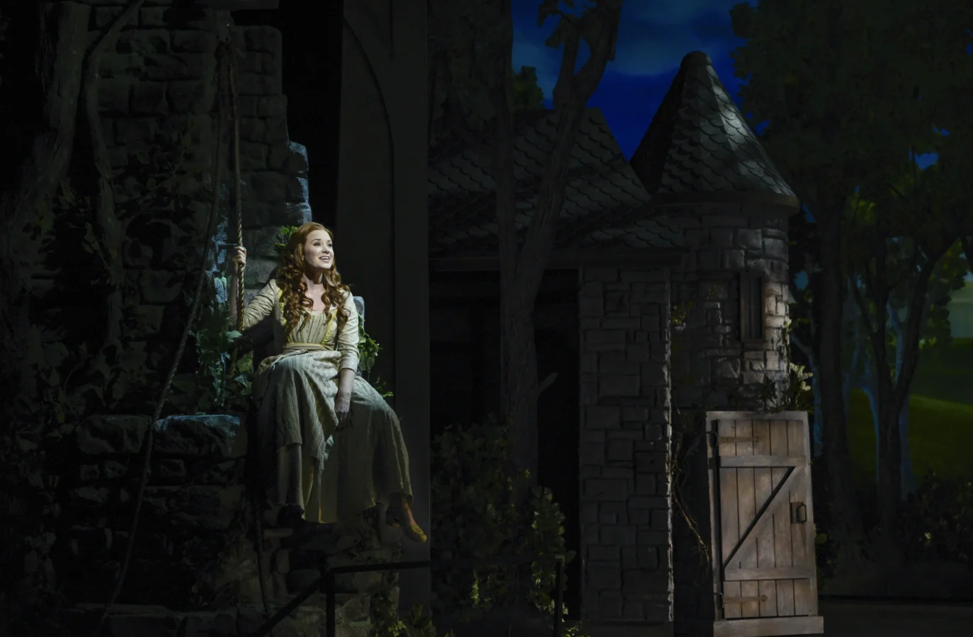 A woman in medieval-style clothing sits on a stone window ledge of a castle set, illuminated by stage lighting, with an open wooden door and trees in the background.