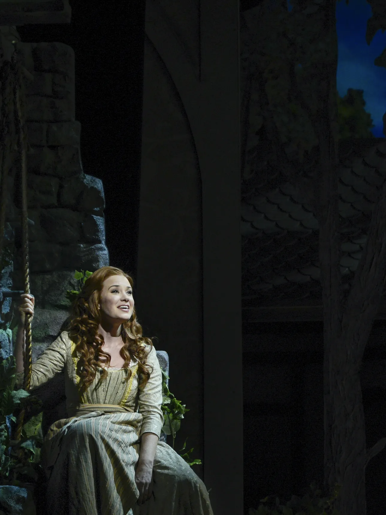 A woman in a long dress sits on a swing, holding a rope and singing on a dimly lit stage with stone and foliage in the background.