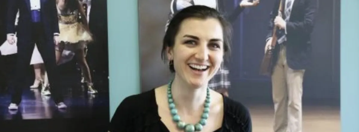 A woman with dark hair and a turquoise beaded necklace smiles in front of posters featuring people in theatrical costumes.
