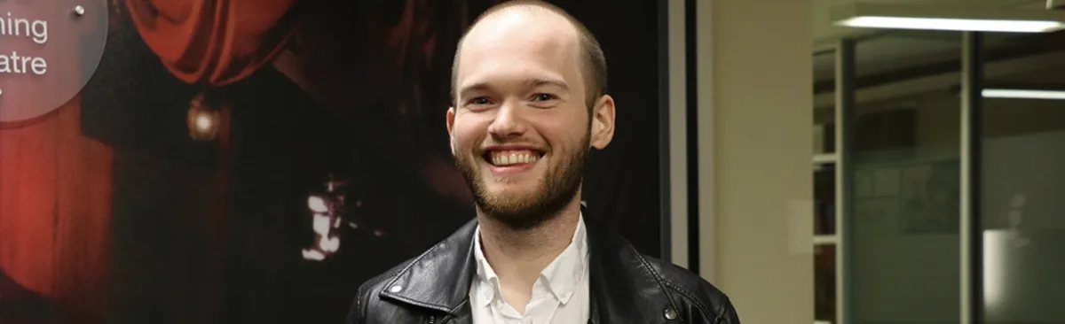 A man with a beard and a shaved head, wearing a black leather jacket and white shirt, smiles while standing indoors near a glass wall.