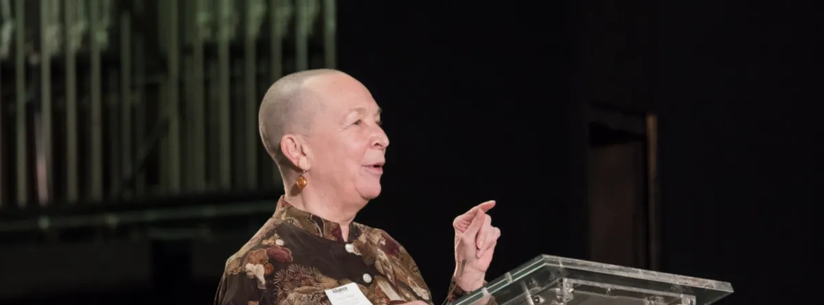 A person with a shaved head speaks at a clear podium, gesturing with one hand. Organ pipes are visible in the blurred background.