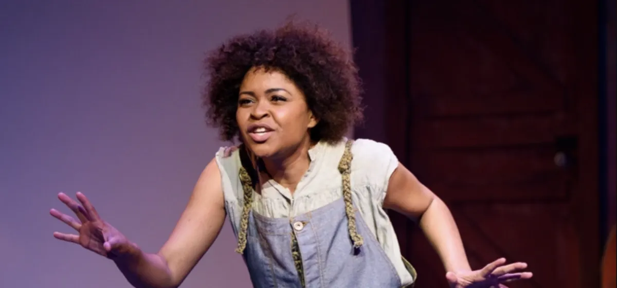 A woman with curly hair stands on stage, wearing a light shirt and overalls, leaning forward with arms outstretched and an expressive facial expression.