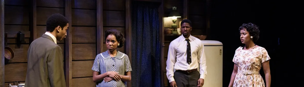 Four people stand in a dimly lit room, dressed in mid-20th century clothing, engaged in a serious conversation near a refrigerator.