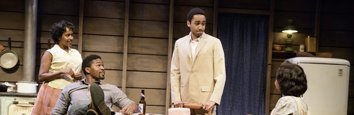 Four people are gathered in a vintage kitchen setting; two are seated at a table, while one man in a light suit stands and a woman stands smiling nearby.