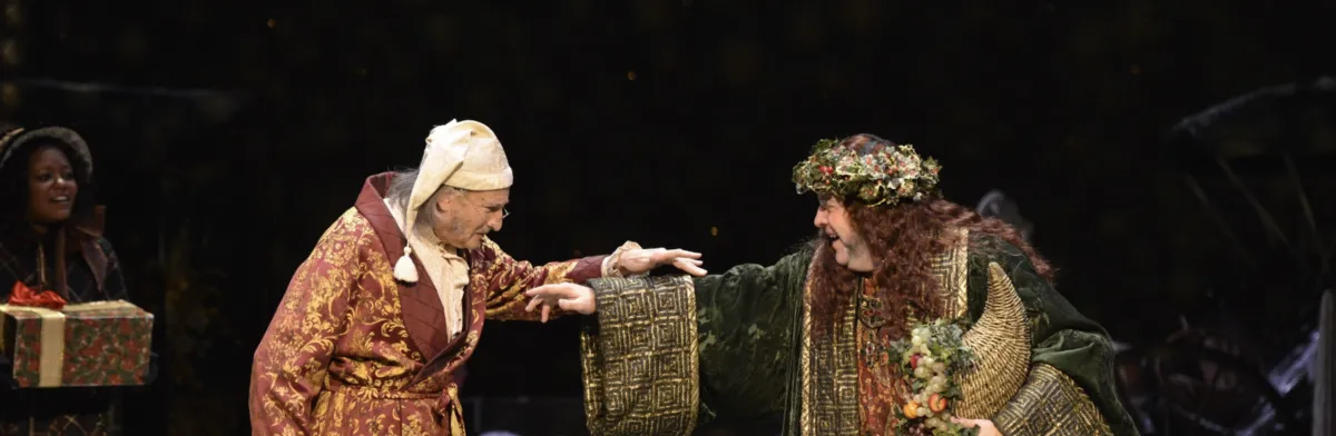 Two actors perform on stage; one is dressed as Ebenezer Scrooge in a nightcap and robe, and the other as the Ghost of Christmas Present in a festive green costume.
