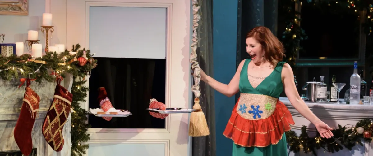 A woman in a green dress and apron holds a curtain aside as food plates appear through a window; festive stockings and decorations are visible.