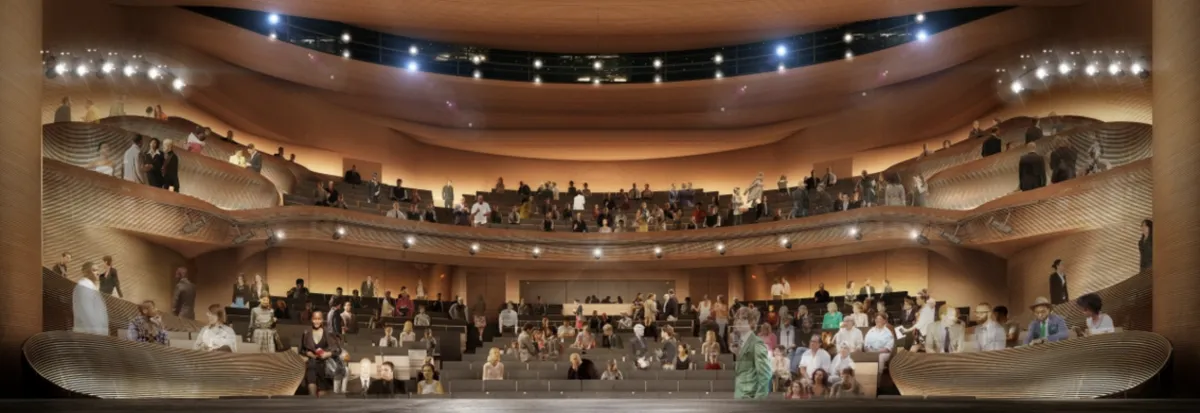 People are seated in multiple tiers of a modern, spacious theater auditorium with warm lighting and curved architectural features.