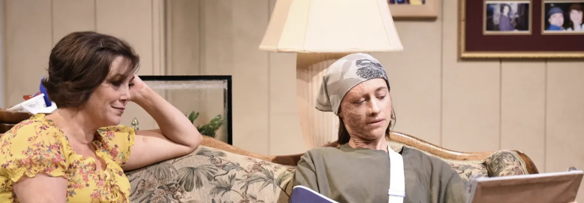 Two women sit on a floral couch; one in a yellow top watches the other, who has facial scars, a bandaged arm, and a head covering, as she looks at a photo album.