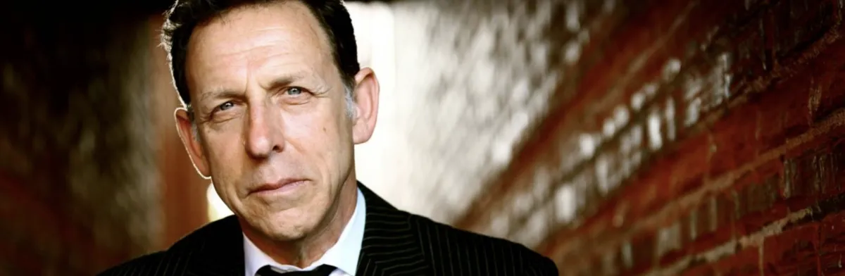 A man in a pinstripe suit and tie stands in a narrow brick alleyway, looking directly at the camera with a neutral expression.
